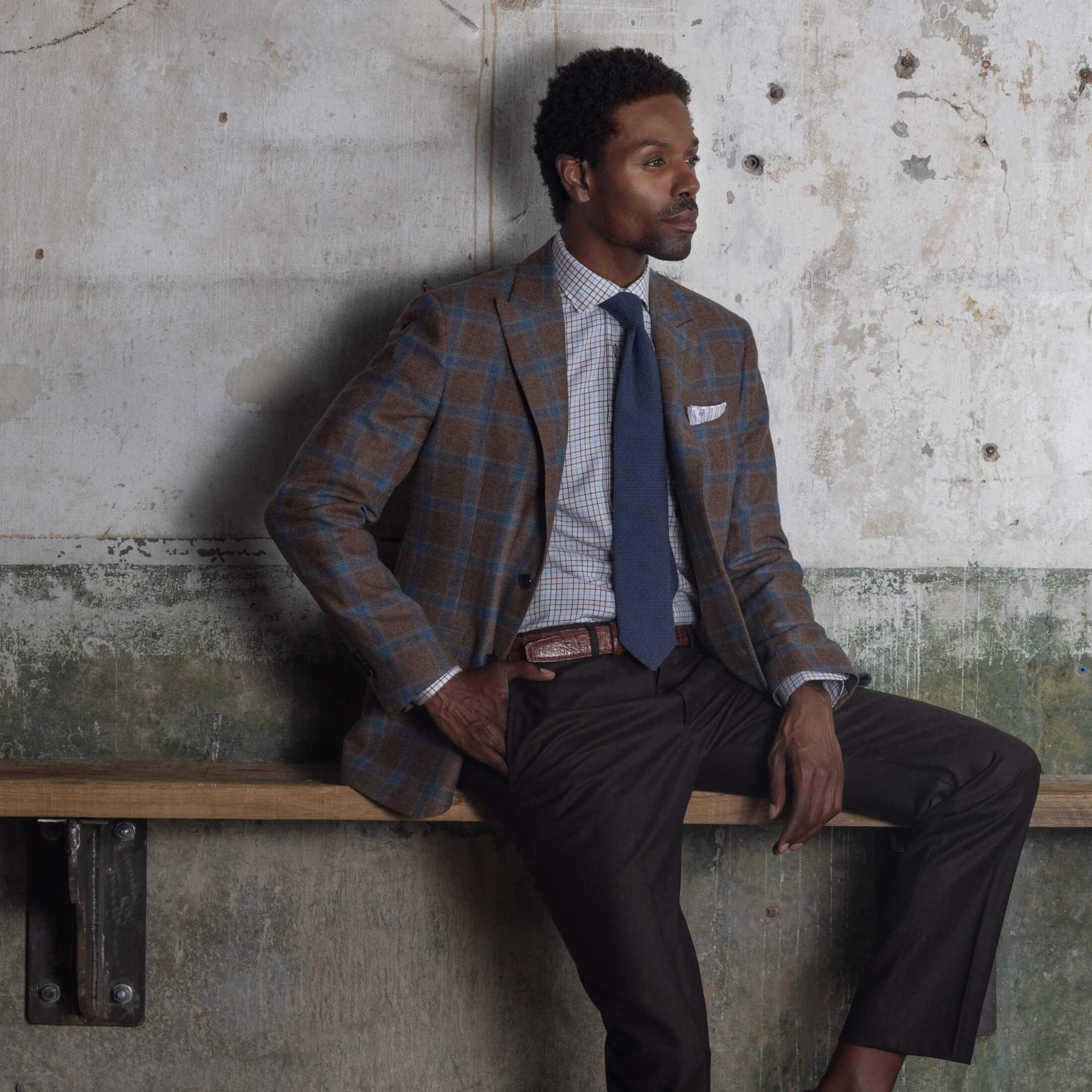 Model sitting on wooden bench against concrete wall wearing Ike Behar sport coat with hand in pocket