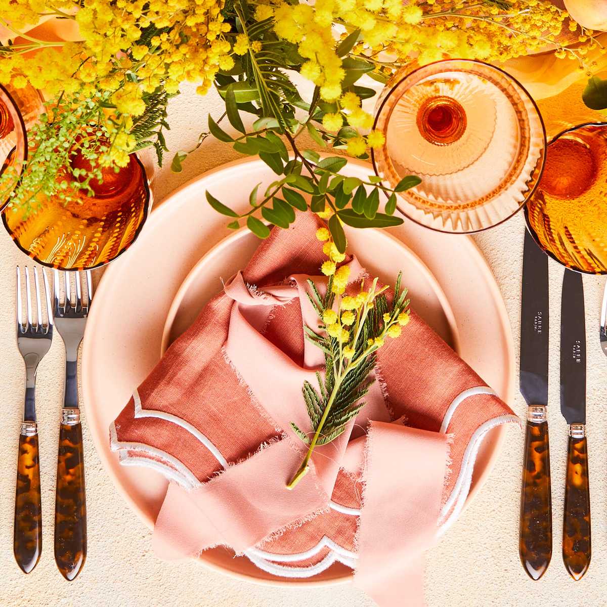 Top view of table setting surrounded by stemware, variety of fruits and flowers on a beige tablecloth